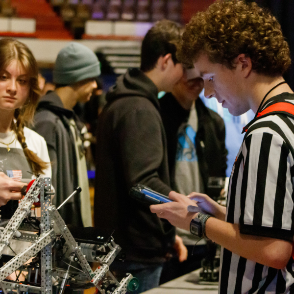 A referee checks a competition robot while students wait during a robotics tournament